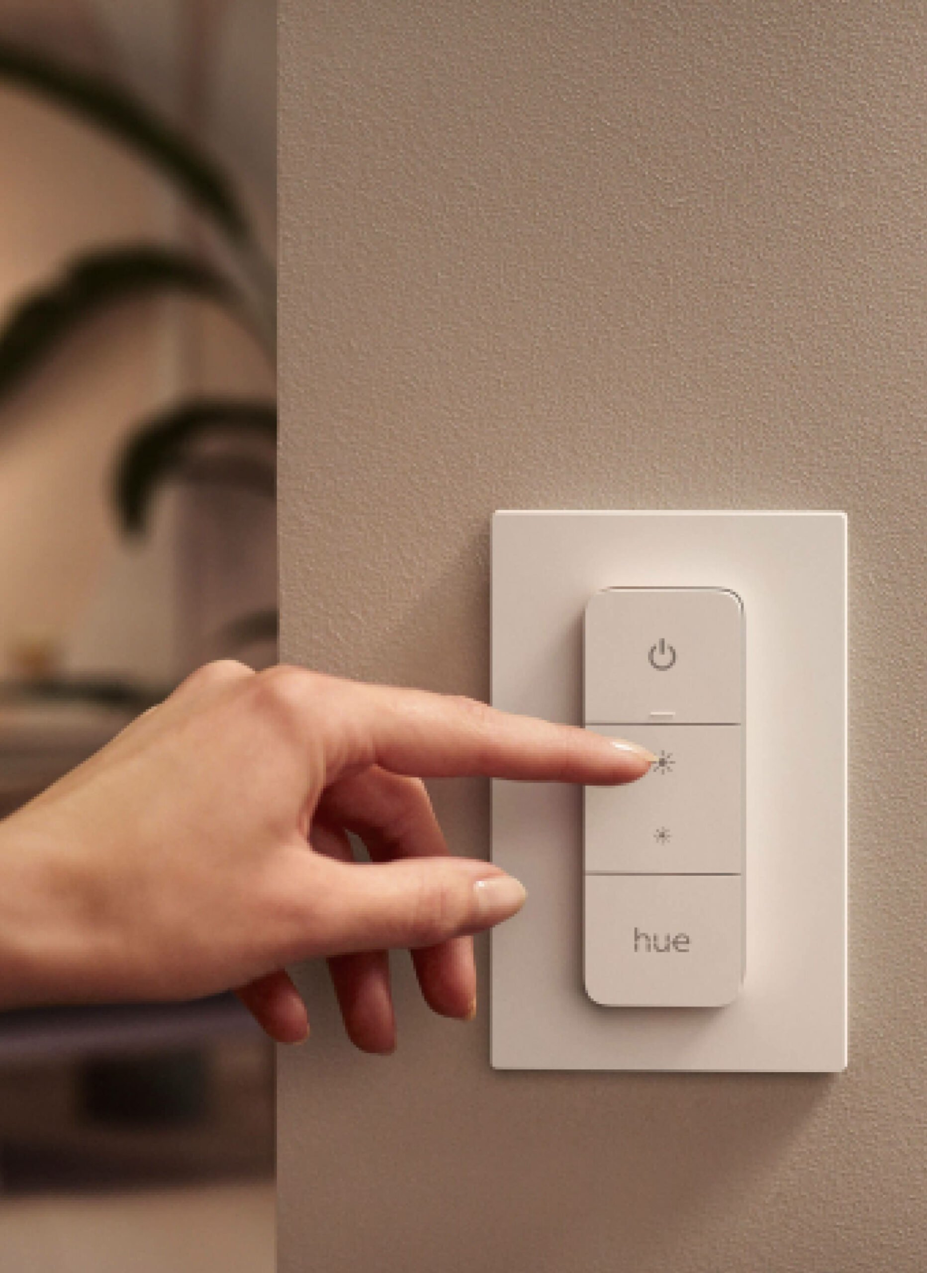 A hand presses the brightness increase button on a wall-mounted Philips Hue light switch.