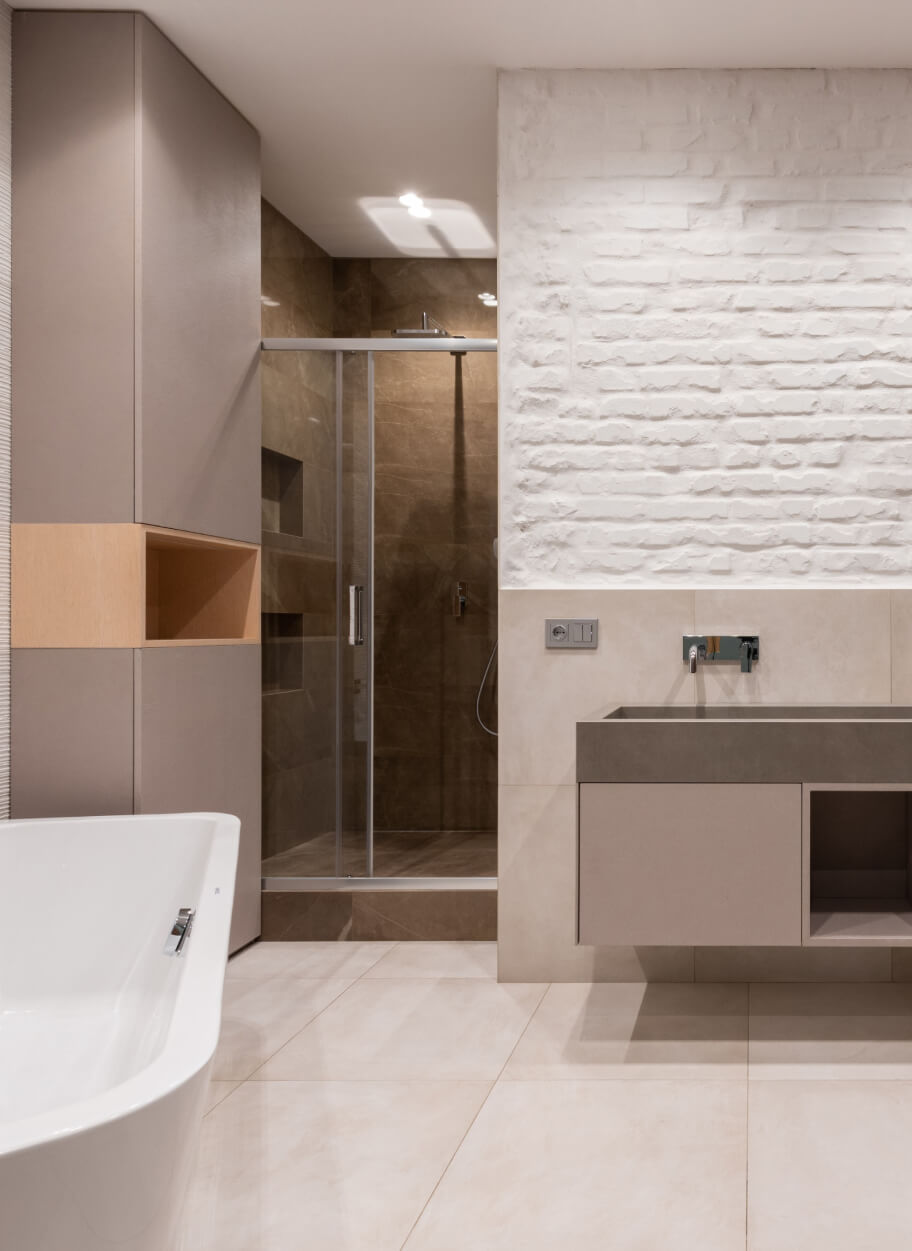 Modern bathroom with a white bathtub, beige vanity, and a glass-enclosed shower with brown tiles, featuring white tile flooring and a white textured brick wall.