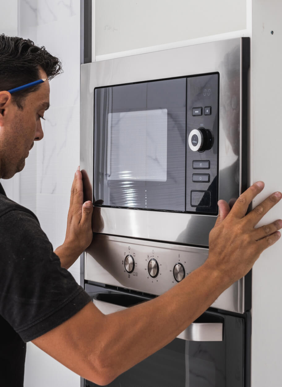 A man installs a built-in microwave above an oven, aligning it with the surrounding cabinetry in a modern kitchen.