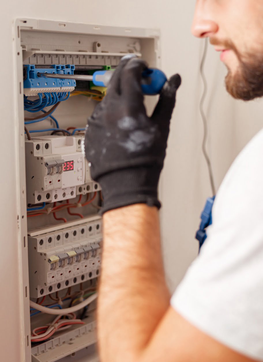A person wearing a black glove uses a screwdriver to work on an open electrical panel with exposed wiring and circuit breakers.