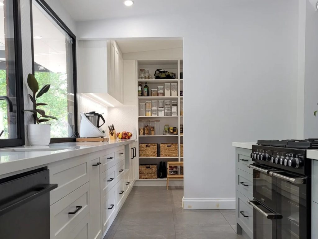 Modern kitchen with white cabinets, black countertop appliances, and a walk-in pantry featuring shelves stocked with baskets, jars, and food items. Bright natural light from a window.