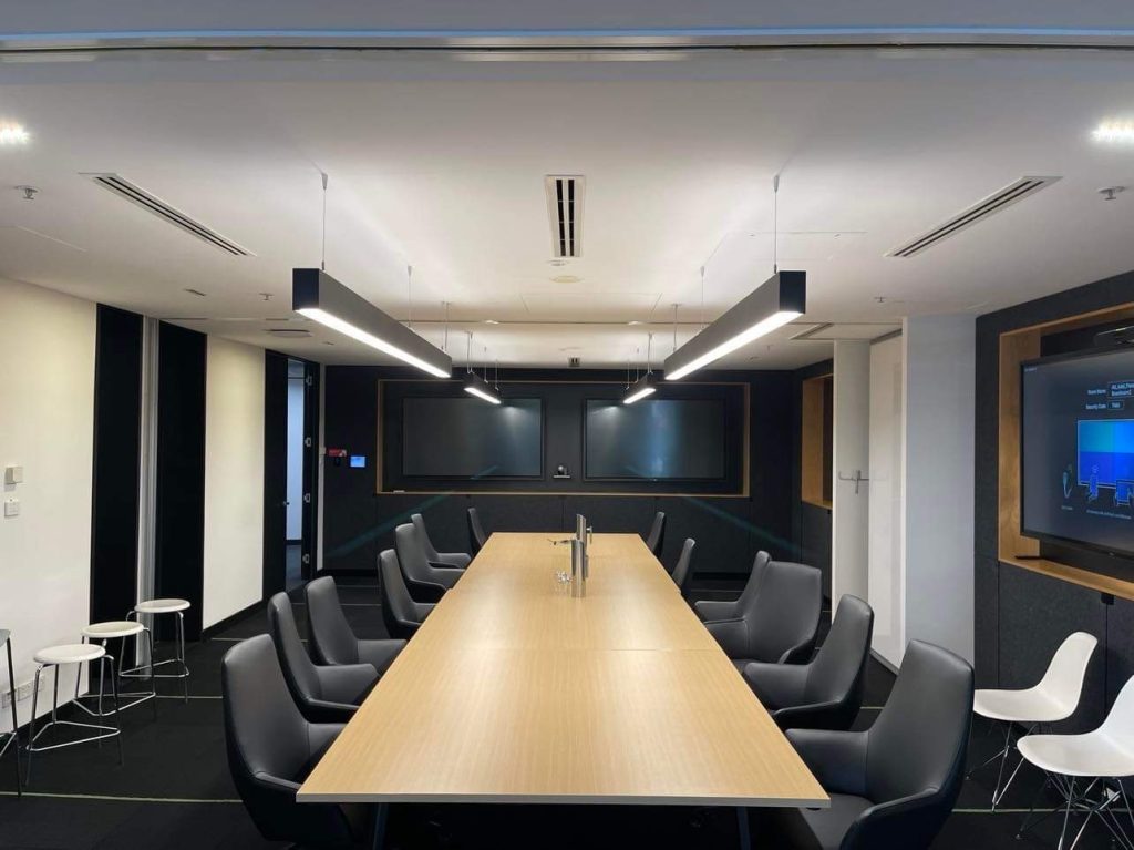 A modern conference room with a long wooden table, black chairs, dual monitors on the far wall, and minimalistic lighting overhead.