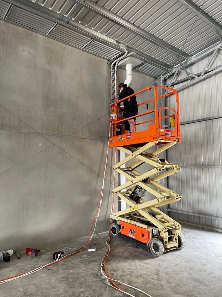 A person stands on an elevated scissor lift, working on electrical cables near the ceiling in an industrial building.