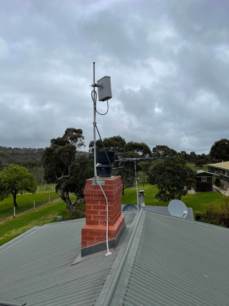 A metal antenna and electronic device are mounted on a brick chimney on a corrugated metal roof, with trees and buildings visible in the background under a cloudy sky.