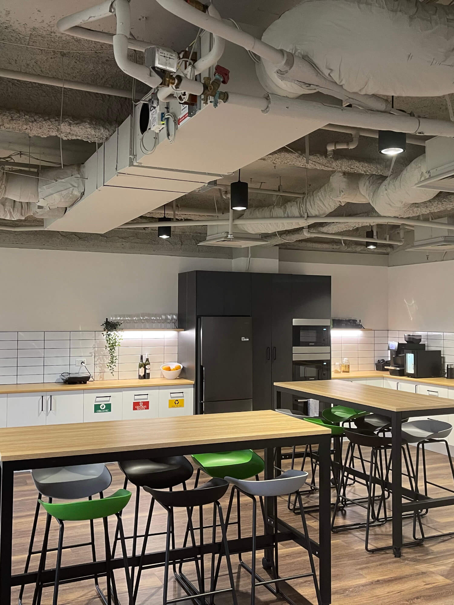 Modern office kitchen with a refrigerator, microwave, and coffee machine, featuring long wooden tables, black and green chairs, and exposed ceiling pipes.
