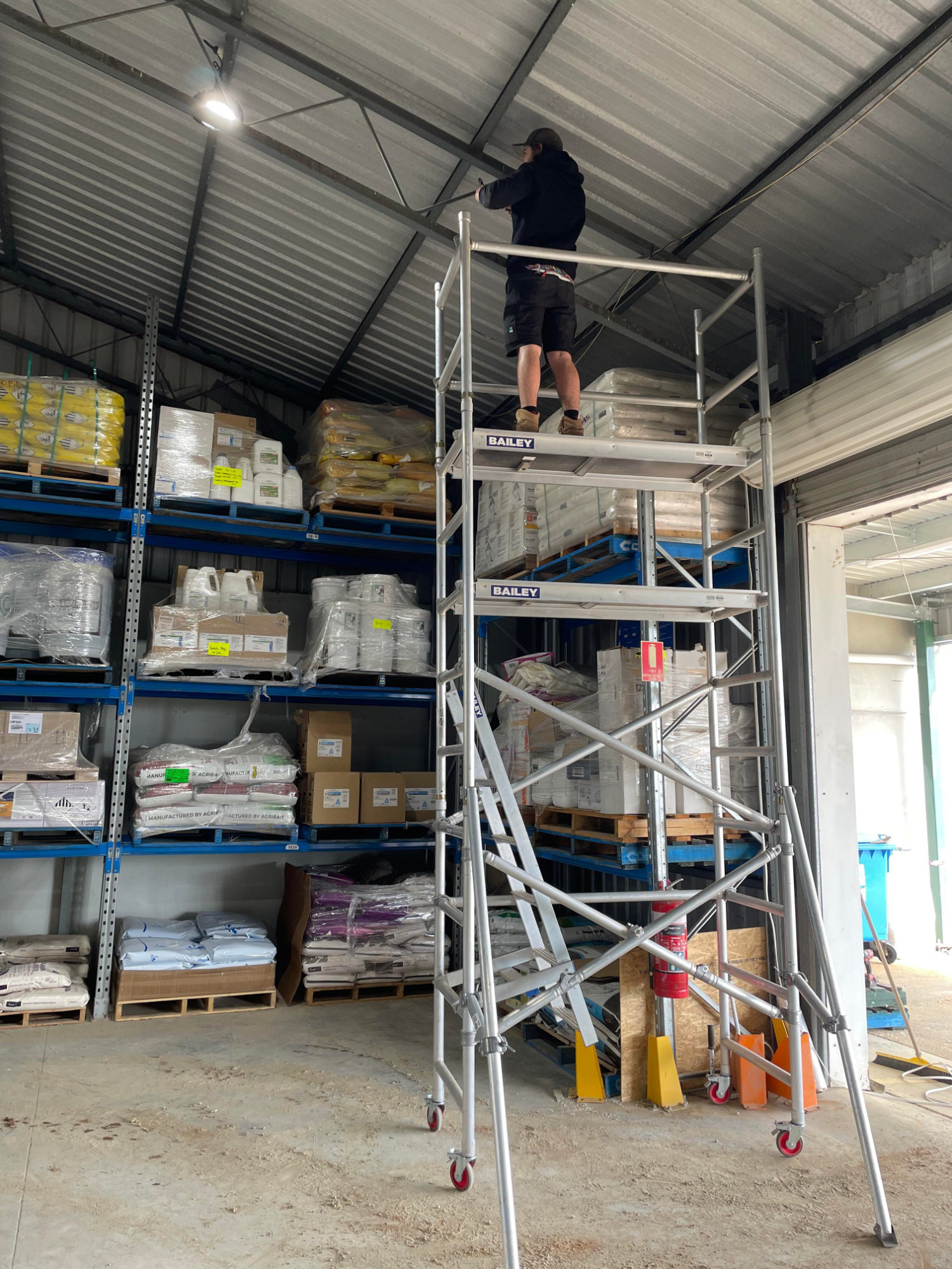 A person stands on a mobile scaffold tower in a warehouse, working near the ceiling. Shelves with stacked goods are visible in the background.