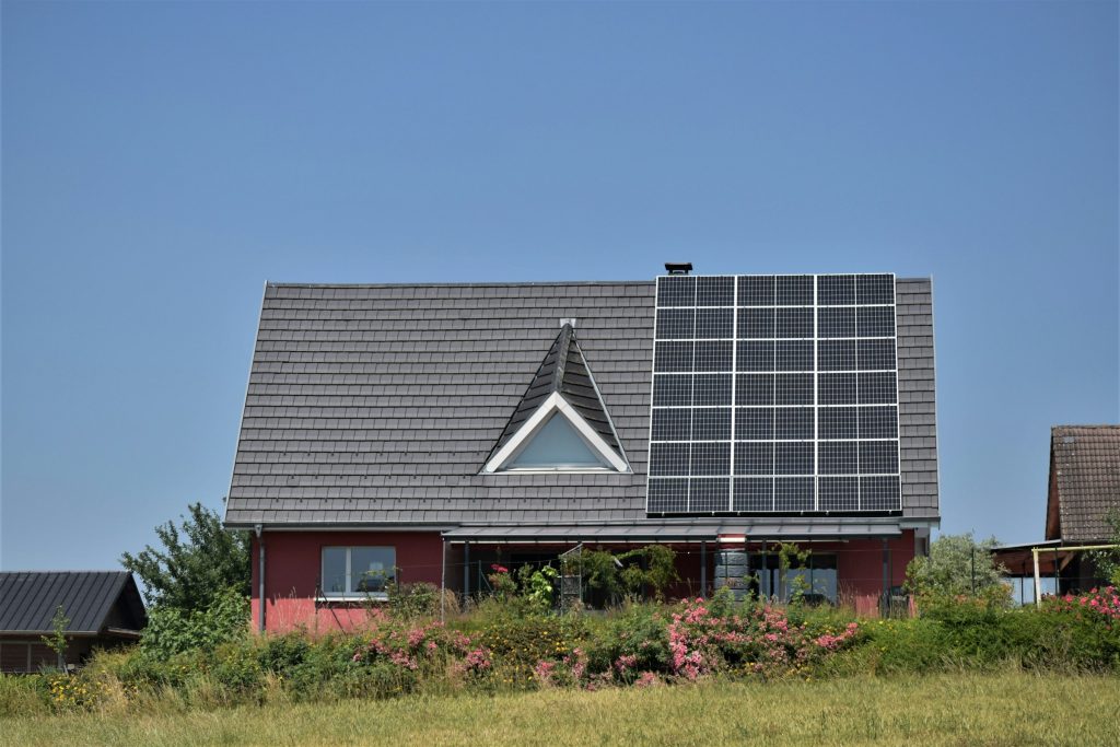 A pink house with a triangular window and Sunpower Maxeon 6 solar panels on the roof, surrounded by lush greenery and vibrant flowers under a clear blue sky.